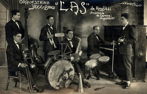 Músics de l'orquestrina jazz-band L'As d'Anglès, sostenint els seus instruments en una sala amb decorat al fons. 1926-1927